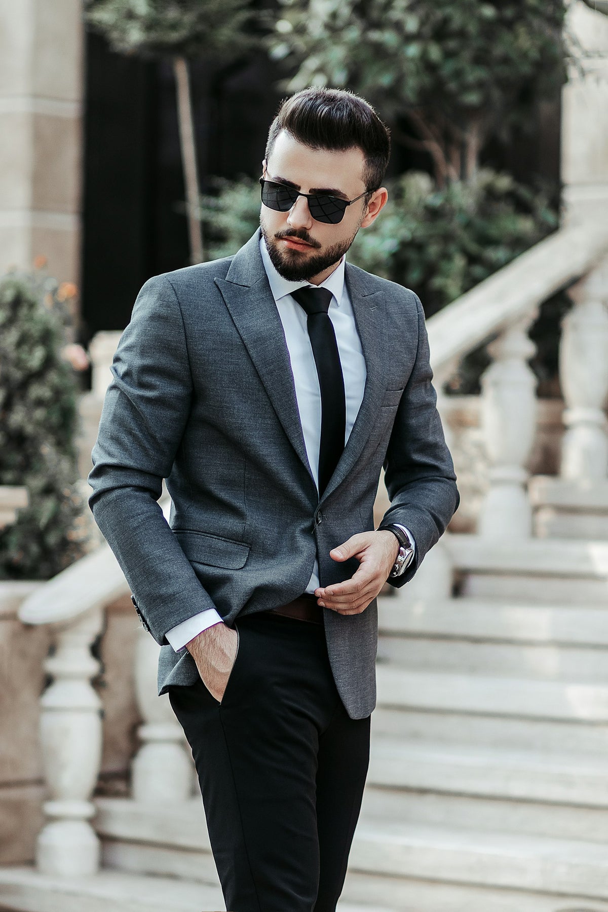 Homem de blazer cinza, calça preta e óculos escuros numa escada, look de esporte fino masculino